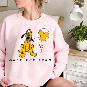 K&ouml;nnte beinhalten: Rosafarbener Pullover mit einem Cartoon-Bild von Pluto, der einen gelben Ballon mit Mickey-Maus-Ohren h&auml;lt. Der Text "Best Day Ever" ist in Schwarz unter dem Bild aufgedruckt.