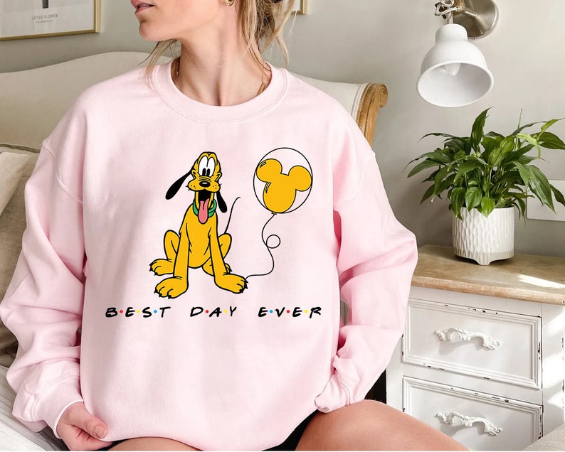 K&ouml;nnte beinhalten: Rosafarbener Pullover mit einem Cartoon-Bild von Pluto, der einen gelben Ballon mit Mickey-Maus-Ohren h&auml;lt. Der Text "Best Day Ever" ist in Schwarz unter dem Bild aufgedruckt.