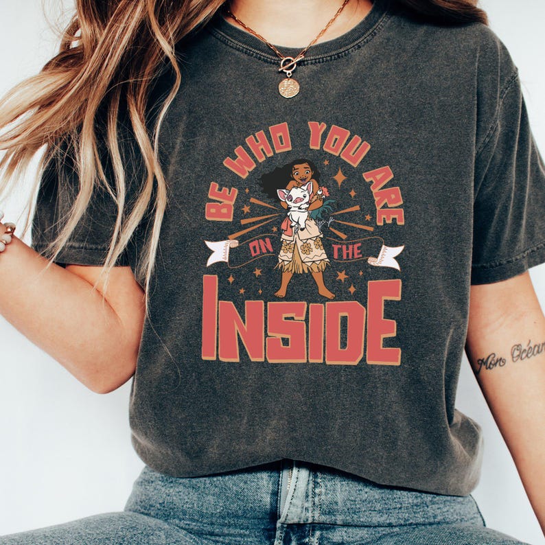 May include: A dark gray t-shirt with a vintage-style graphic featuring a cartoon character holding a small white animal. The text on the graphic reads "Be who you are on the inside".