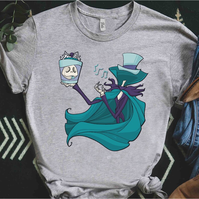 Puede incluir: Una camiseta gris con una ilustraci&oacute;n en turquesa y morado de un personaje de la pel&iacute;cula Pesadilla antes de Navidad. El personaje sostiene una caja con una calavera dentro y canta.