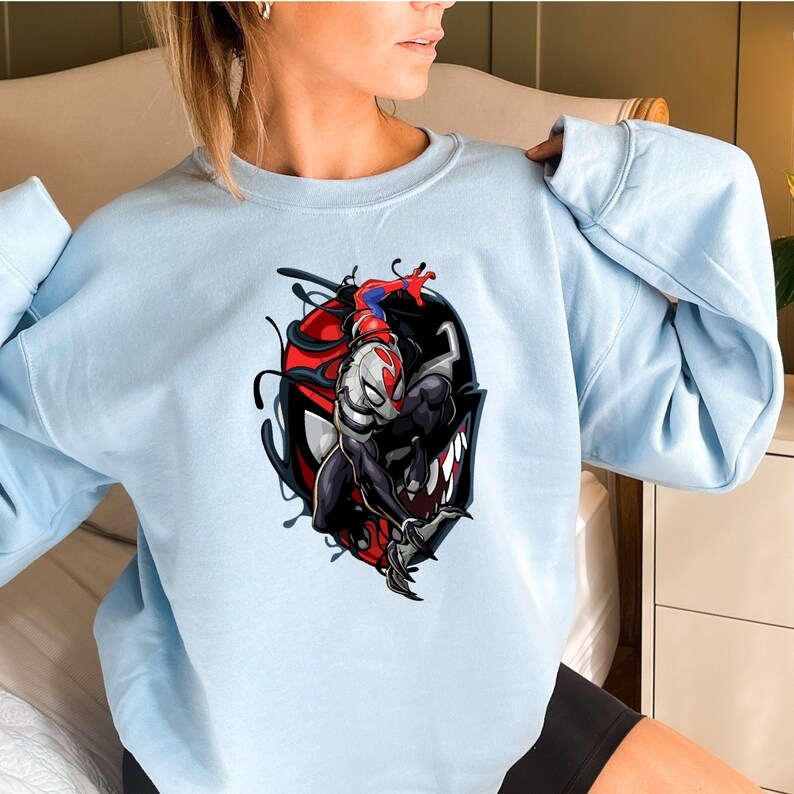 Puede incluir: Una sudadera de color azul claro con un estampado gr&aacute;fico de un personaje de superh&eacute;roe negro y rojo con una m&aacute;scara blanca. El personaje est&aacute; en una pose de lucha con los brazos levantados.