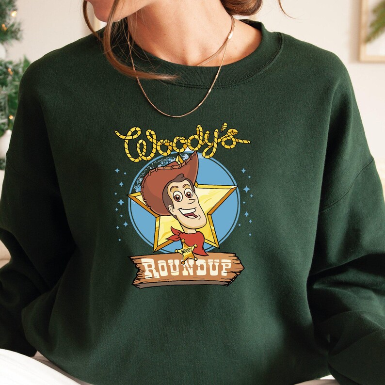Peut inclure: Un sweat-shirt vert fonc&eacute; avec une image de dessin anim&eacute; de Woody de Toy Story. L'image comprend le texte "Woody's Roundup".