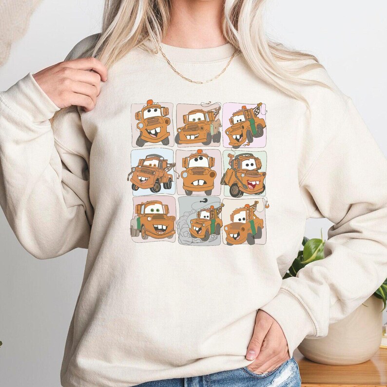 Puede incluir: Una sudadera beige con una imagen de dibujos animados de una gr&uacute;a de remolque con diferentes expresiones faciales en una cuadr&iacute;cula.