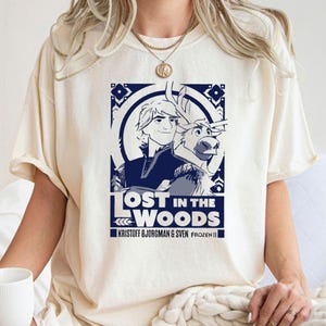 Puede incluir: Una camiseta blanca con un gr&aacute;fico azul de Kristoff y Sven de Frozen II. El gr&aacute;fico dice "Lost in the Woods" y "Kristoff Bjorgman & Sven Frozen II".