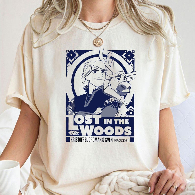Puede incluir: Una camiseta blanca con un gr&aacute;fico azul de Kristoff y Sven de Frozen II. El gr&aacute;fico dice "Lost in the Woods" y "Kristoff Bjorgman & Sven Frozen II".