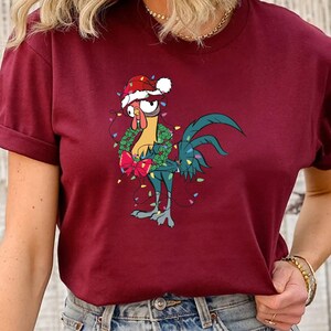 May include: A maroon t-shirt with a cartoon rooster wearing a Santa hat and a Christmas wreath around its neck. The rooster is surrounded by Christmas lights.