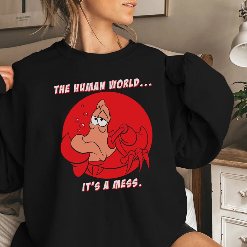 Op de afbeelding: Zwart sweatshirt met een rode cirkel en een cartoonkrab erin. De tekst "THE HUMAN WORLD... IT'S A MESS" staat op het sweatshirt gedrukt.