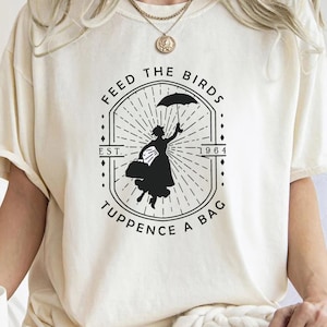 May include: White t-shirt with a black and white graphic design of a woman holding an umbrella and a bag. The graphic says "Feed the Birds Est. 1964 Tuppence a Bag".