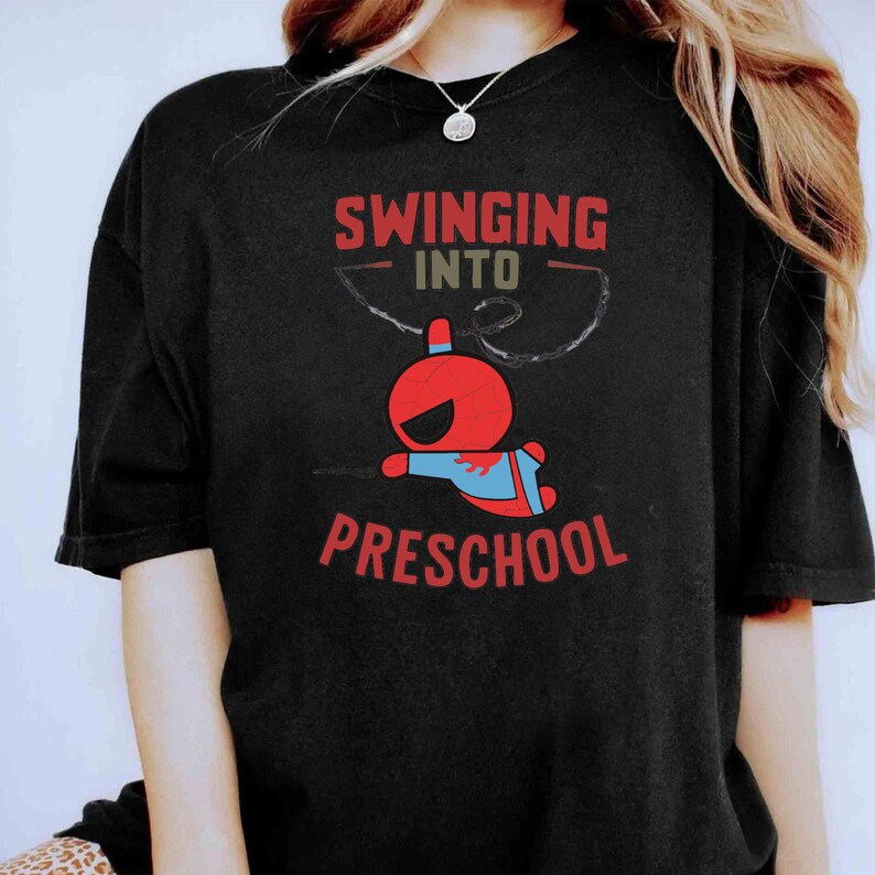 Puede incluir: Camiseta negra con un personaje de dibujos animados rojo y azul balance&aacute;ndose en una telara&ntilde;a y el texto "Swinging into Preschool".