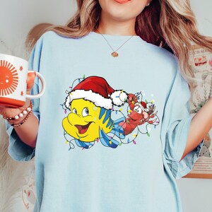 Puede incluir: Una camiseta azul claro con un gr&aacute;fico navide&ntilde;o de Flounder de La Sirenita con un gorro de Pap&aacute; Noel y Sebasti&aacute;n el cangrejo sosteniendo luces navide&ntilde;as.
