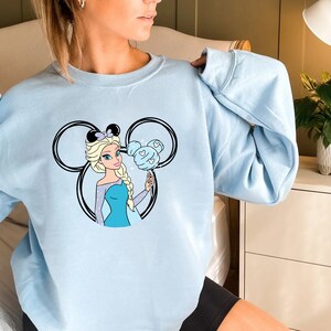 Puede incluir: Sudadera azul claro con un gr&aacute;fico de una princesa de dibujos animados sosteniendo un algod&oacute;n de az&uacute;car azul en forma de oreja de Mickey Mouse.