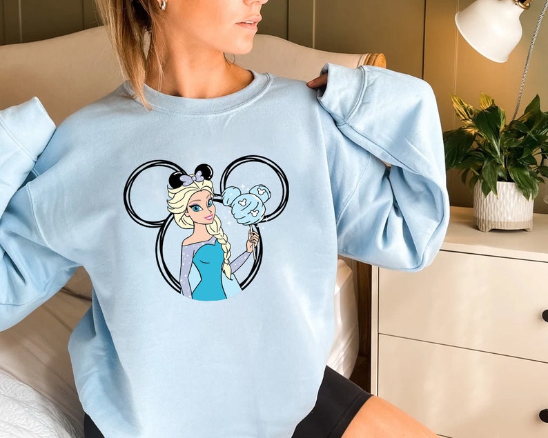 Puede incluir: Sudadera azul claro con un gr&aacute;fico de una princesa de dibujos animados sosteniendo un algod&oacute;n de az&uacute;car azul en forma de oreja de Mickey Mouse.