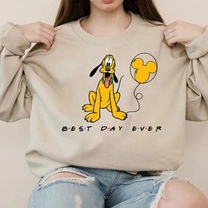 K&ouml;nnte beinhalten: Ein beiger Pullover mit einem Cartoon-Bild von Pluto, der einen gelben Ballon mit einem Mickey-Mouse-Ohren-Design h&auml;lt. Der Text "BEST DAY EVER" ist unter dem Bild aufgedruckt.