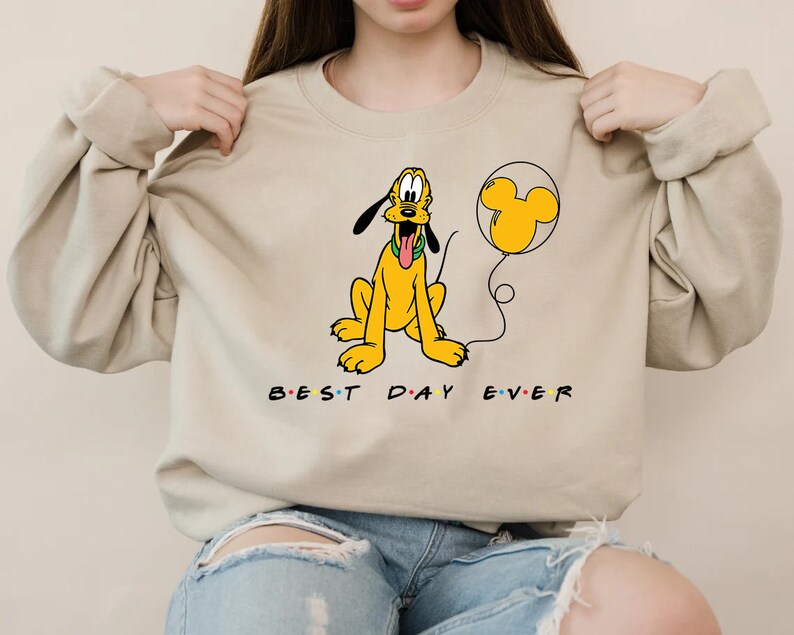 K&ouml;nnte beinhalten: Ein beiger Pullover mit einem Cartoon-Bild von Pluto, der einen gelben Ballon mit einem Mickey-Mouse-Ohren-Design h&auml;lt. Der Text "BEST DAY EVER" ist unter dem Bild aufgedruckt.