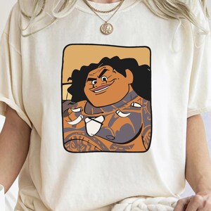 May include: A white t-shirt featuring a cartoon illustration of a Polynesian man with brown skin and black hair. The man is wearing a brown and grey patterned shirt and has a mischievous expression on his face.