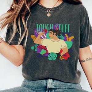 May include: Dark gray t-shirt with the text "TOUGH STUFF" in green. The graphic features a woman flexing her arm, surrounded by colorful flowers and butterflies. The shirt is a comfortable, casual style.