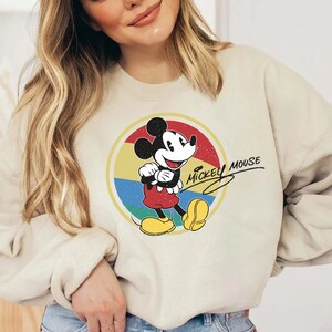 Puede incluir: Una sudadera de color crema con un gr&aacute;fico de Mickey Mouse de estilo vintage. El gr&aacute;fico presenta a Mickey Mouse con un atuendo rojo y amarillo, de pie en un c&iacute;rculo con un fondo de arco&iacute;ris. El texto "Mickey Mouse" est&aacute; escrito en una fuente cursiva sobre Mickey Mouse.