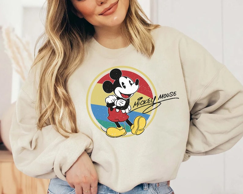 Puede incluir: Una sudadera de color crema con un gr&aacute;fico de Mickey Mouse de estilo vintage. El gr&aacute;fico presenta a Mickey Mouse con un atuendo rojo y amarillo, de pie en un c&iacute;rculo con un fondo de arco&iacute;ris. El texto "Mickey Mouse" est&aacute; escrito en una fuente cursiva sobre Mickey Mouse.
