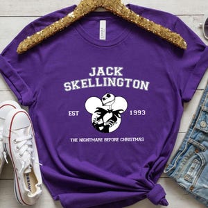 May include: Purple t-shirt with a white graphic of Jack Skellington from "The Nightmare Before Christmas" in a Mickey Mouse silhouette. The text "Jack Skellington" is above the graphic, and "EST 1993" is below the graphic.