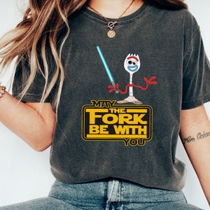 K&ouml;nnte beinhalten: Ein dunkelgraues T-Shirt mit einem Cartoon-Bild von Forky aus Toy Story, der ein Lichtschwert h&auml;lt. Der Text "M&ouml;ge die Gabel mit dir sein" ist in Gelb auf schwarzem Hintergrund gedruckt.