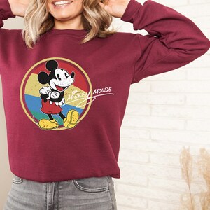 Puede incluir: Una sudadera color burdeos con un gr&aacute;fico de estilo vintage de Mickey Mouse en un c&iacute;rculo amarillo, azul y verde. El texto "Mickey Mouse" est&aacute; escrito en cursiva blanca sobre el c&iacute;rculo.