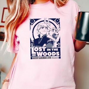 Puede incluir: Una camiseta rosa con un estampado gr&aacute;fico de Kristoff y Sven de Frozen II. El gr&aacute;fico presenta una ilustraci&oacute;n azul y blanca de Kristoff y Sven con el texto "Lost in the Woods" y "Kristoff Bjorgman & Sven Frozen II".