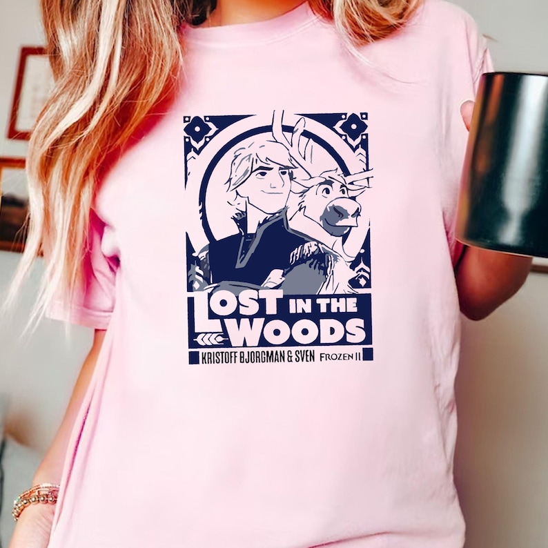 Puede incluir: Una camiseta rosa con un estampado gr&aacute;fico de Kristoff y Sven de Frozen II. El gr&aacute;fico presenta una ilustraci&oacute;n azul y blanca de Kristoff y Sven con el texto "Lost in the Woods" y "Kristoff Bjorgman & Sven Frozen II".