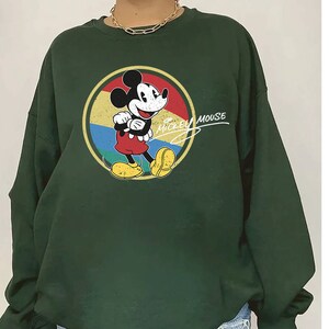 Puede incluir: Una sudadera verde oscuro con un gr&aacute;fico vintage de Mickey Mouse en un c&iacute;rculo colorido. El texto "Mickey Mouse" est&aacute; escrito en cursiva blanca sobre el gr&aacute;fico.