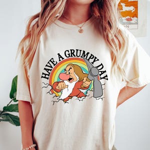 May include: A white t-shirt featuring a graphic of Grumpy from Snow White holding a pickaxe. The graphic is surrounded by a rainbow and clouds. The text "Have a Grumpy Day" is written around the graphic.