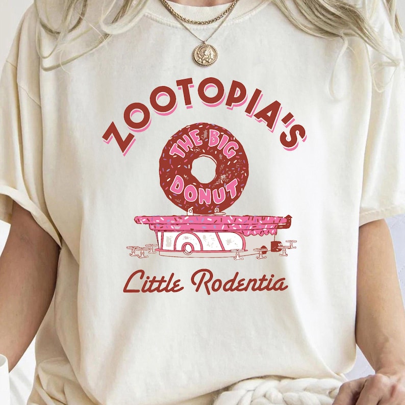 May include: A white t-shirt with a pink and red graphic design that says "Zootopia's The Big Donut Little Rodentia". The graphic features a large donut with pink sprinkles.