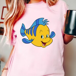 May include: A pink t-shirt with a cartoon yellow fish with blue fins and a blue fin on its head. The fish has a smiling face with a large mouth and small eyes.