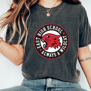 May include: A gray t-shirt with a red and white circle graphic that says "East High School Always A Wildcat". The graphic features a red wildcat head.