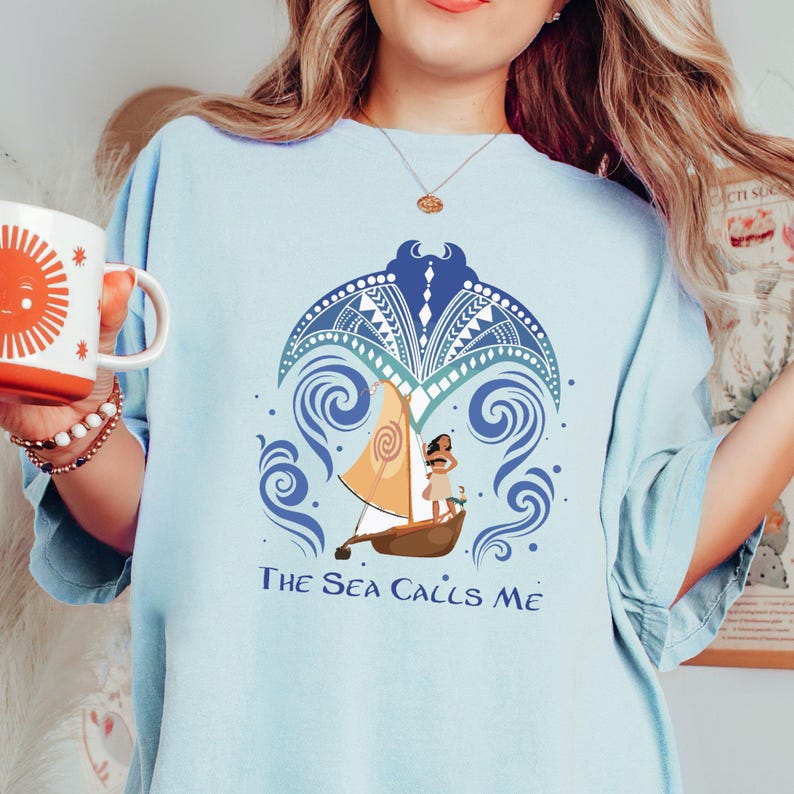May include: A light blue t-shirt with a graphic of a Polynesian woman sailing on a boat with the text "The Sea Calls Me".