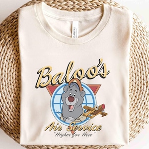 May include: A beige t-shirt with a cartoon illustration of Baloo from Disney's Jungle Book. The illustration features Baloo wearing a pilot's hat and holding a plane. The text "Baloo's Air Service" and "Higher for Hire" is printed on the shirt.