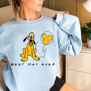 K&ouml;nnte beinhalten: Hellblauer Pullover mit einem Cartoon-Bild von Pluto, der einen Mickey-Maus-Ballon h&auml;lt. Der Text "BEST DAY EVER" ist unter dem Bild aufgedruckt.