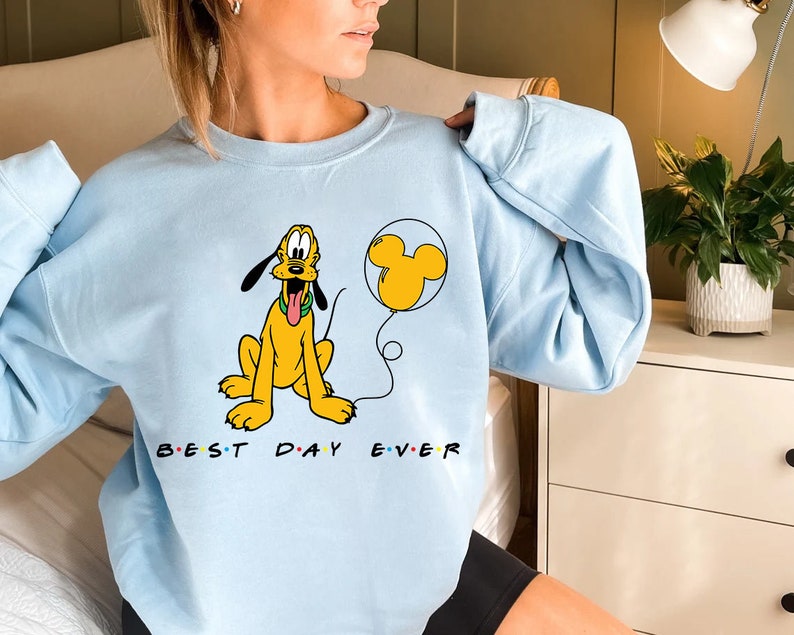 K&ouml;nnte beinhalten: Hellblauer Pullover mit einem Cartoon-Bild von Pluto, der einen Mickey-Maus-Ballon h&auml;lt. Der Text "BEST DAY EVER" ist unter dem Bild aufgedruckt.