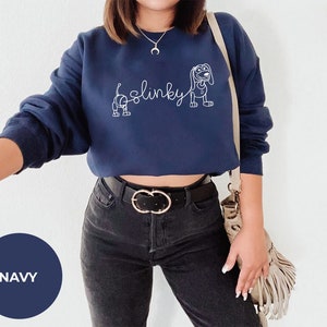 May include: A navy blue cropped sweatshirt with a white outline of Slinky Dog from the Toy Story franchise. The sweatshirt has the word "Slinky" written in cursive.