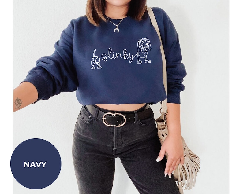 May include: A navy blue cropped sweatshirt with a white outline of Slinky Dog from the Toy Story franchise. The sweatshirt has the word "Slinky" written in cursive.