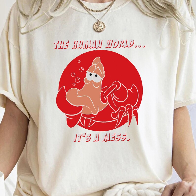 Op de afbeelding: Een wit T-shirt met een rode cirkel en een cartoonkrab erin. De tekst "THE HUMAN WORLD... IT'S A MESS" staat op het shirt gedrukt.
