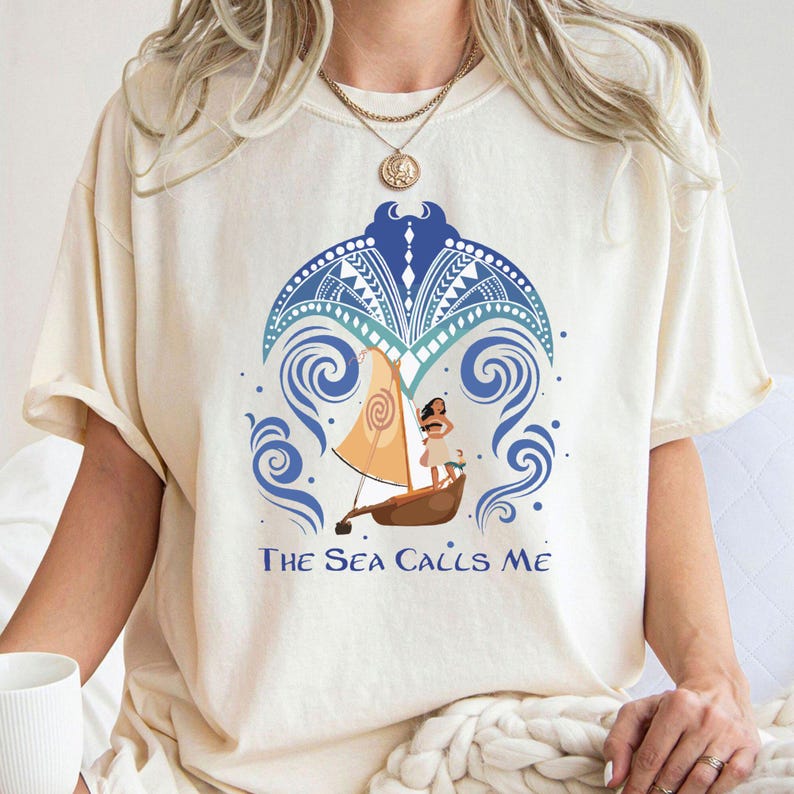 May include: A white t-shirt with a blue and white design of a Polynesian woman sailing on a boat with the text "The Sea Calls Me".