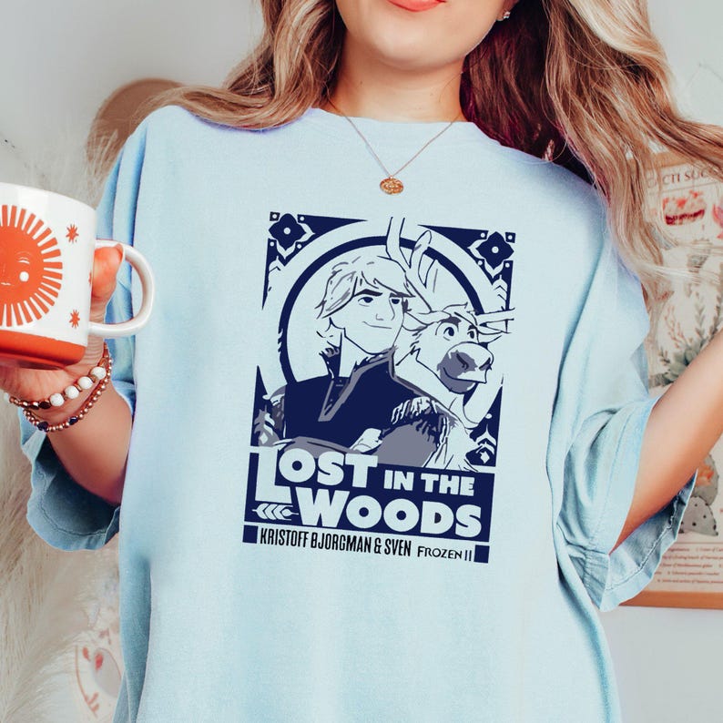 Puede incluir: Una camiseta azul claro con un gr&aacute;fico de Kristoff y Sven de Frozen II. El gr&aacute;fico es de estilo vintage con el texto "Lost in the Woods" y "Kristoff Bjorgman & Sven Frozen II".