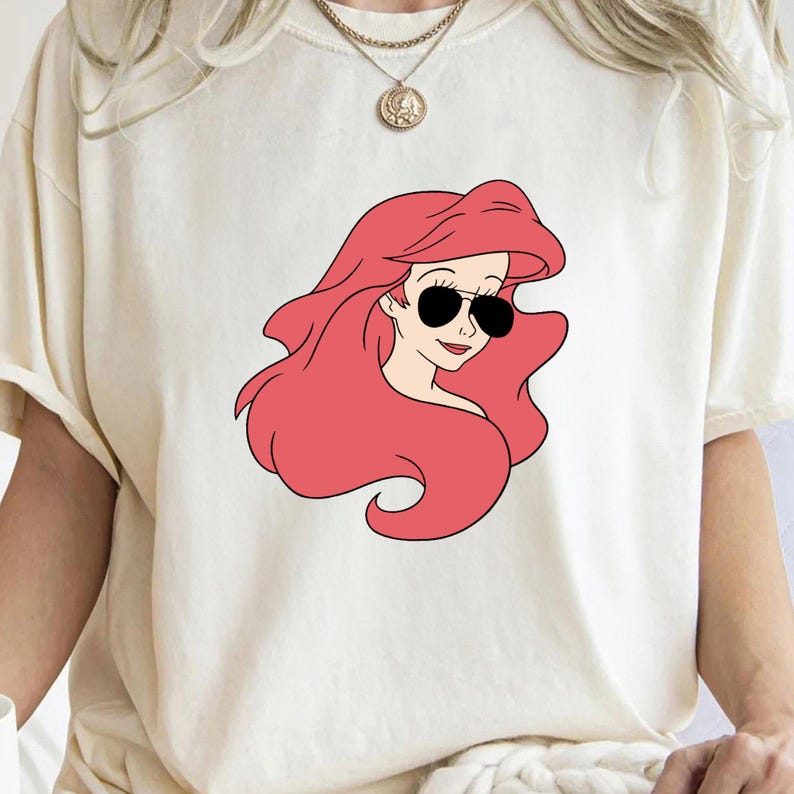 May include: A white t-shirt with a red-haired cartoon character wearing sunglasses. The character is a mermaid with a pink tail.