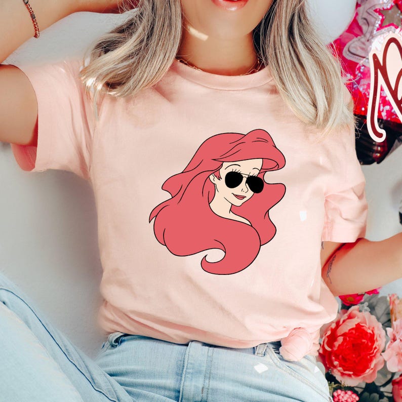 May include: Pink t-shirt with a cartoon image of Ariel from The Little Mermaid wearing sunglasses.
