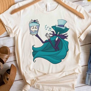 Puede incluir: Una camiseta blanca con una ilustraci&oacute;n de dibujos animados de un personaje que lleva un sombrero de copa azul y una capa turquesa que sostiene un frasco de vidrio con una calavera en su interior. Notas musicales flotan sobre la cabeza del personaje.