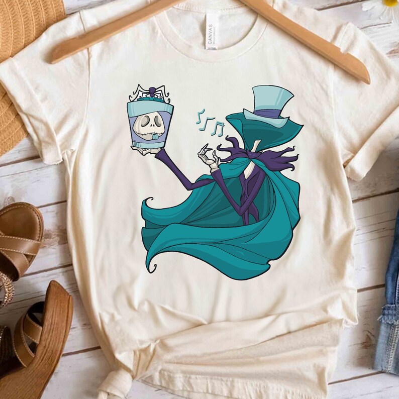Puede incluir: Una camiseta blanca con una ilustraci&oacute;n de dibujos animados de un personaje que lleva un sombrero de copa azul y una capa turquesa que sostiene un frasco de vidrio con una calavera en su interior. Notas musicales flotan sobre la cabeza del personaje.