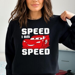 Puede incluir: Sudadera negra con un coche de carreras rojo de dibujos animados y el texto "Speed I am Speed" en blanco.