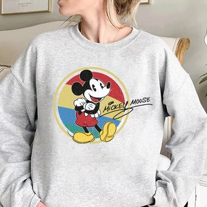 Puede incluir: Una sudadera gris con un gr&aacute;fico de estilo vintage de Mickey Mouse en un c&iacute;rculo rojo y amarillo. El texto "Mickey Mouse" est&aacute; escrito en cursiva negra.