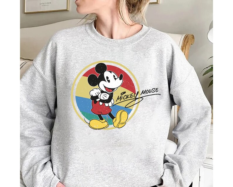 Puede incluir: Una sudadera gris con un gr&aacute;fico de estilo vintage de Mickey Mouse en un c&iacute;rculo rojo y amarillo. El texto "Mickey Mouse" est&aacute; escrito en cursiva negra.