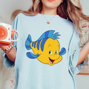 May include: A light blue t-shirt with a cartoon image of a yellow and blue fish with a smiling face. The fish is a character from the Disney movie The Little Mermaid.