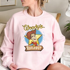 Peut inclure: Sweat-shirt rose avec une image de dessin anim&eacute; de Woody de Toy Story. Le graphique repr&eacute;sente le visage de Woody et le texte "Woody's Roundup".